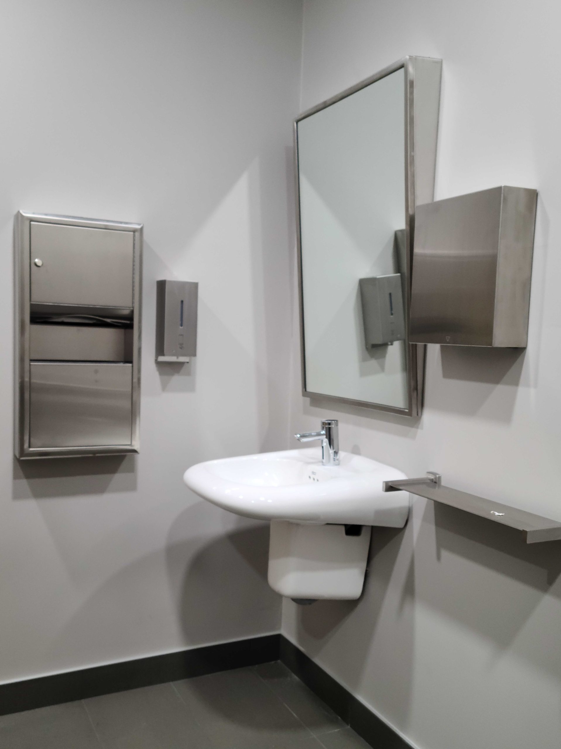 Low restroom sink with downward-angled mirror, dispensers and receptacles for toiletries, and a small shelf
