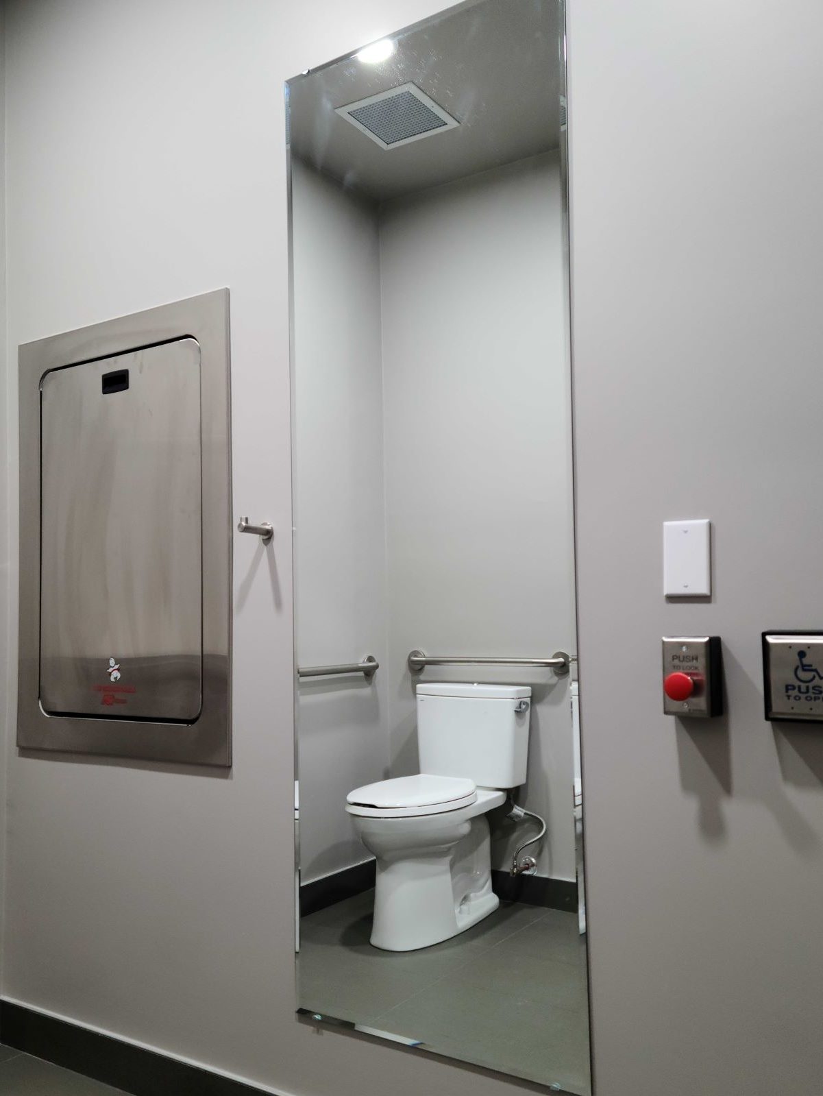 Restroom wall featuring changing table, clothing hook, full-length mirror, and large buttons for opening or locking the door
