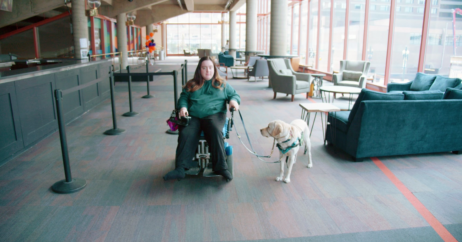 Carly Neis and her service animal, Gilmore, in the Shoctor lobby. We see the Shoctor lobby bar and seating area in the background. Carly is a light skinned person with long brown hair who uses a powered wheelchair and Gilmore is a light coloured, medium sized dog.