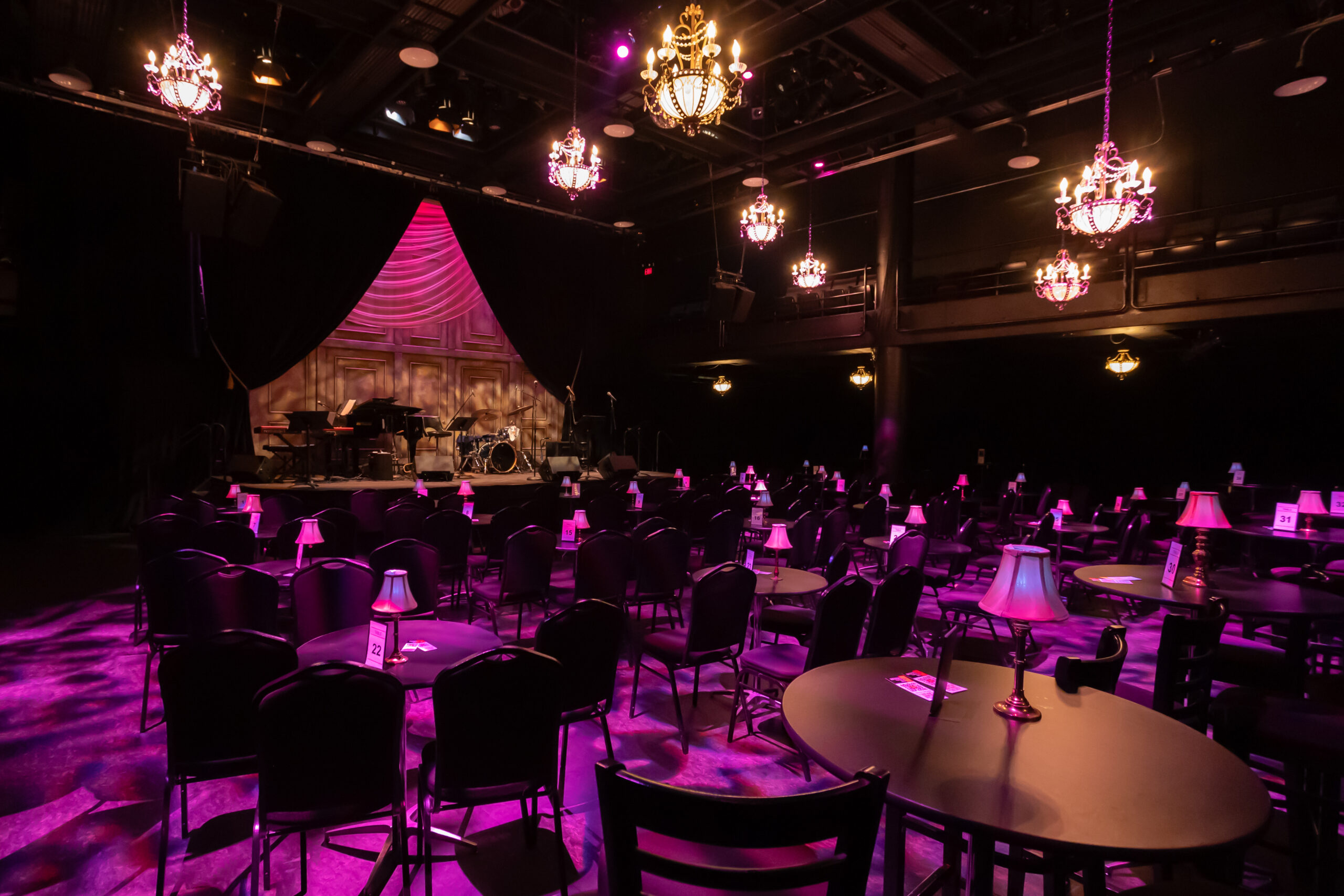 A photograph of the Rice Theatre when set as a cabaret. The venue has many tables and chairs. The tables have small lamps on them and chandeliers hang above the seating area. The Rice Theatre stage is shown with instruments.